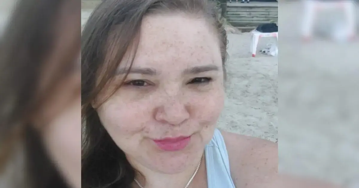 Pessoa sorrindo à beira da praia, com cabelo solto e pele clara, em um dia ensolarado.