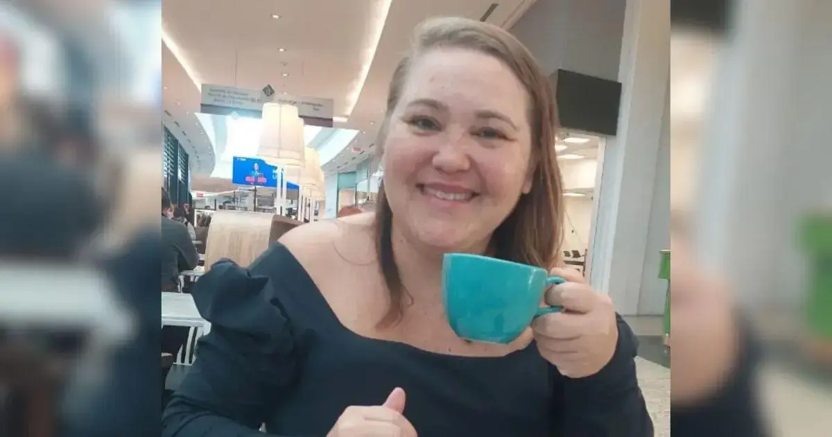 Mulher sorrindo em um café, segurando uma caneca azul e sentada em uma mesa.