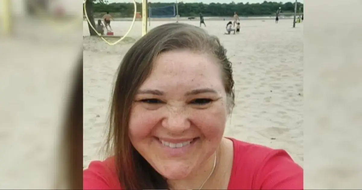 Pessoa sorrindo em uma praia com areia clara, ao fundo, outras pessoas brincando e praticando atividades.