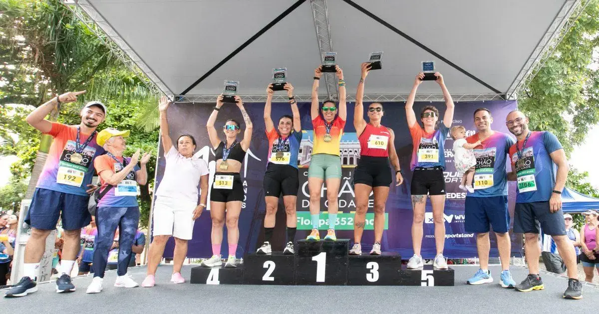 Pódio da corrida com participantes segurando troféus e medalhas, cercados por pessoas e bandeiras do evento.