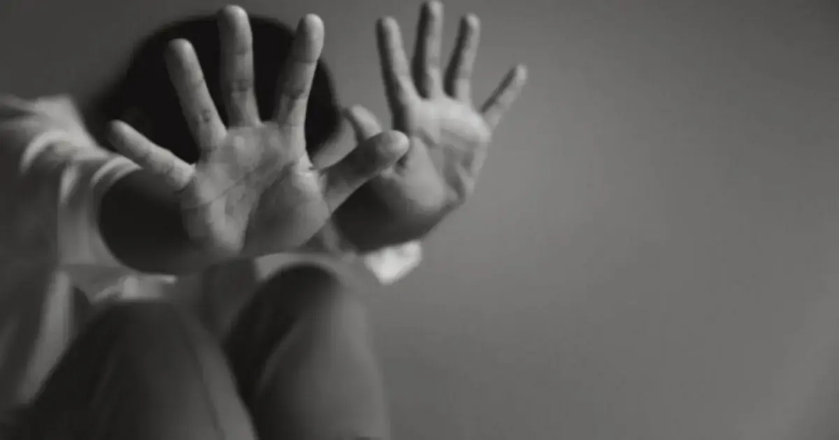 Mãos abertas em gesto de defesa, com uma pessoa sentada no fundo, em um ambiente com iluminação suave.