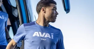 Atleta em uniforme azul do Avaí saindo de um ônibus, com a palavra "AVAI" visível em destaque.