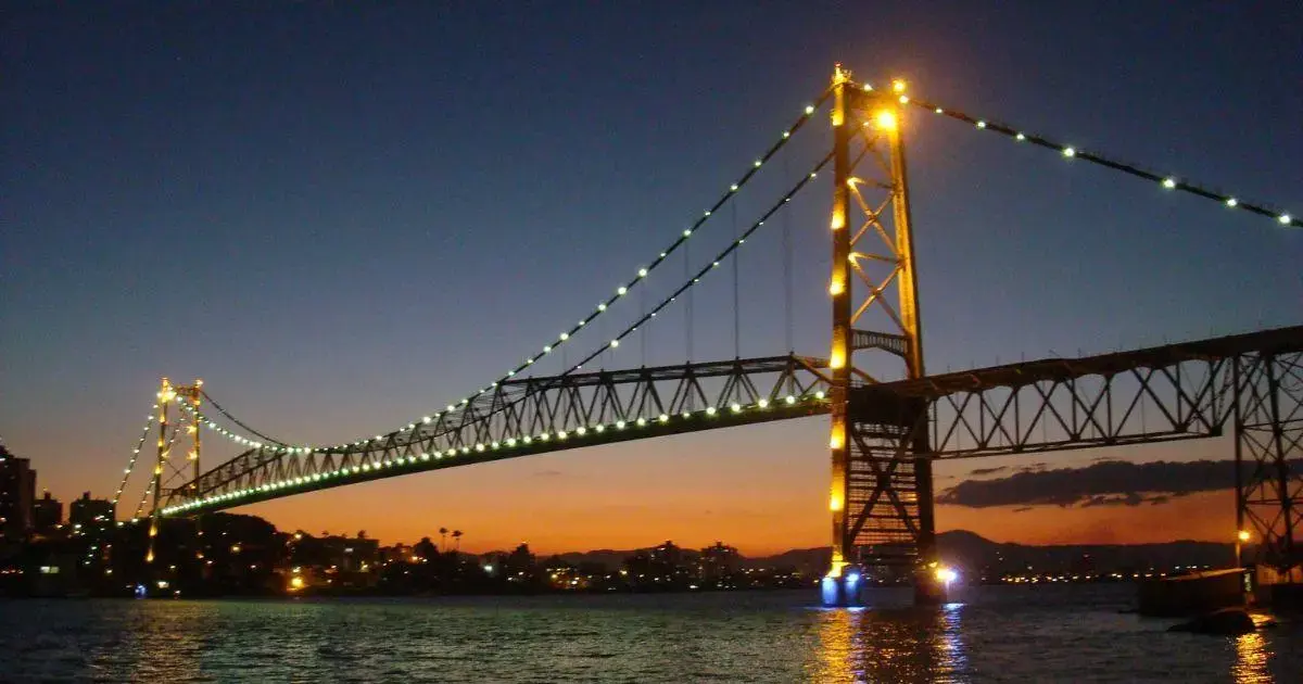 Ponte iluminada à noite, com céu gradualmente escurecendo e reflexos na água.