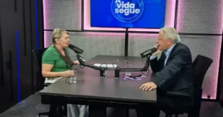Duas pessoas sentadas em uma mesa de estúdio, conversando com microfones e copos de água. No fundo, um painel azul com o texto "A vida segue".