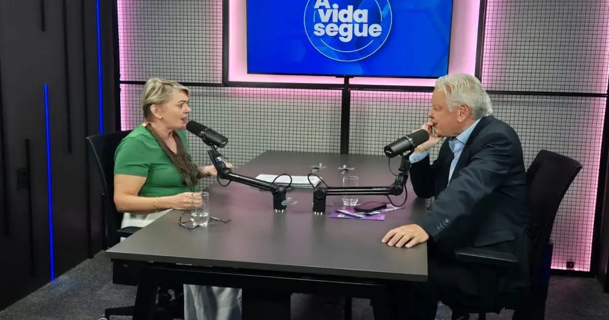 Duas pessoas sentadas em uma mesa de estúdio, conversando com microfones e copos de água. No fundo, um painel azul com o texto 