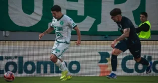Jogadores em campo durante uma partida de futebol, com bandeira ao fundo e torcida visível.