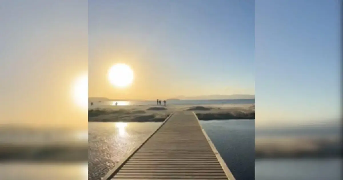 Deck de madeira sobre um rio com pessoas ao fundo admirando o pôr do sol à beira da praia.