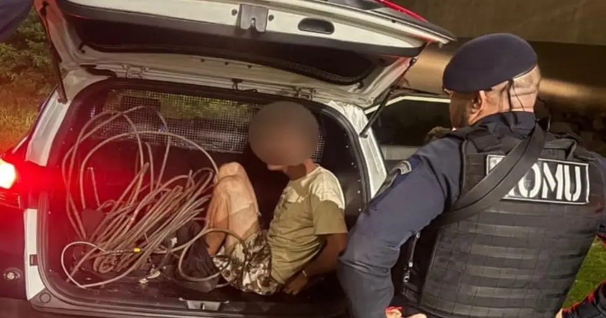 Homem sentado no porta-malas de um veículo com fios ao seu redor, acompanhado por um agente de segurança.