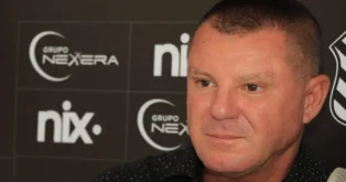 Homem com cabelo curto e camisa escura, com painéis de patrocinadores ao fundo.