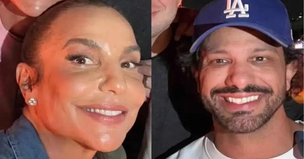 Duas pessoas sorrindo, uma com brincos e a outra usando um boné azul, posando juntas em um ambiente iluminado.