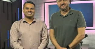 Dois homens sorrindo em um estúdio com fundo iluminado em tons de rosa e azul.