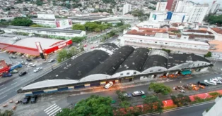 Vista aérea de um prédio com telhado arredondado, cercado por ruas, veículos e árvores em uma área urbana.