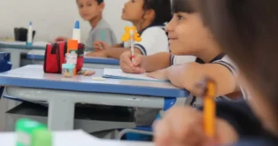 Crianças em sala de aula, concentradas em atividade, com materiais escolares dispostos nas mesas.