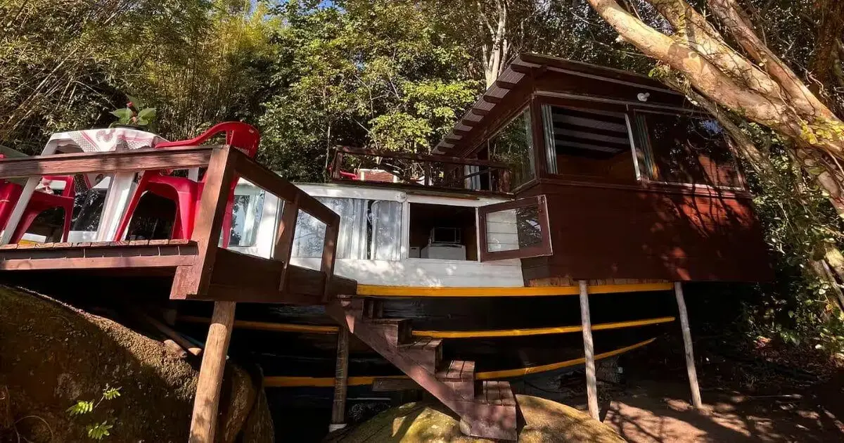 Casa-barco em meio à vegetação, com escada de acesso, mesas e cadeiras vermelhas ao redor.