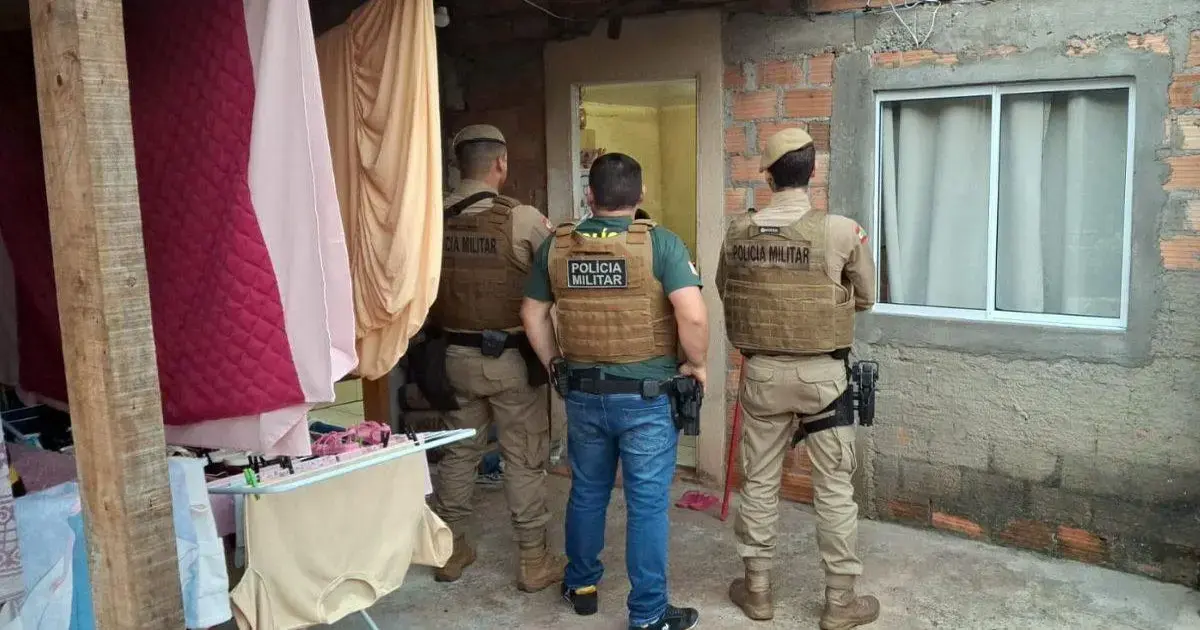 Três agentes da Polícia Militar estão em frente a uma entrada de imóvel, observando o interior.