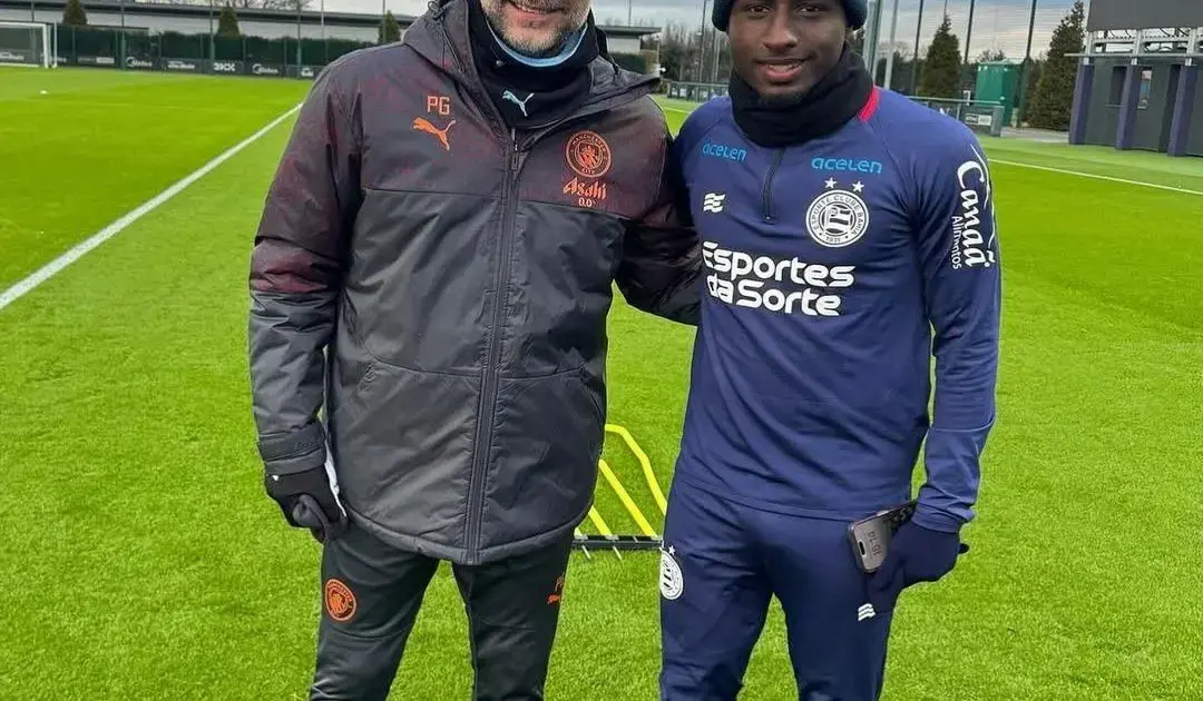 Dois homens sorrindo em um campo de futebol, um vestindo jaqueta preta e o outro em uniforme azul.