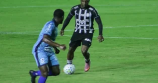 Jogadores em campo durante uma partida de futebol, um atleta com uniforme preto driblando um adversário com uniforme azul.