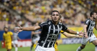 Jogador em campo, comemorando um gol com os braços abertos, enquanto a torcida ao fundo observa.