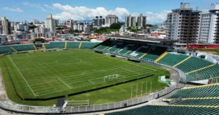 Estádio com campo de futebol verde, arquibancadas e prédios ao fundo sob um céu claro.