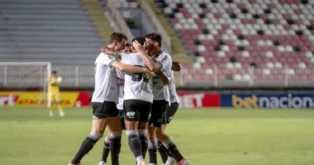Jogadores celebram juntos após um gol, em campo, com arquibancadas vazias ao fundo.