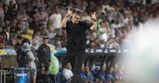 Treinador faz sinal de positivo enquanto chove durante uma partida, com torcedores ao fundo.