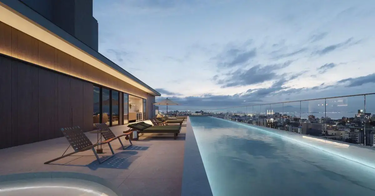 Pôr do sol visto de uma piscina infinita em um terraço com espreguiçadeiras e uma cidade ao fundo.