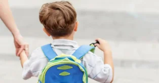 Criança de costas segurando uma mochila colorida, enquanto é acompanhada por um adulto, em ambiente urbano.