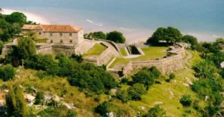 Construção histórica com paredes de pedra, cercada por vegetação, localizada à beira de uma praia.