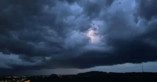 Céu escuro com nuvens carregadas e relâmpago visível ao fundo, iluminação na área da cidade.