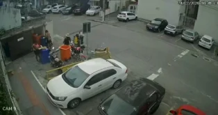 Duas pessoas em frente a lixeiras coloridas, uma moto ao lado e carros estacionados na rua.