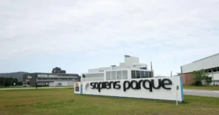 Sinal de entrada do Sapiens Parque em Florianópolis, com edifícios de vidro e gramado ao redor.