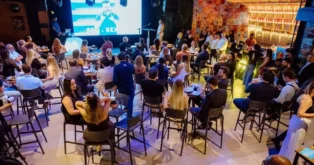 Pessoas sentadas em mesas, assistindo a uma apresentação em um evento em um local iluminado.