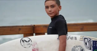 Criança com roupa de surfe preta segurando pranchas de surfe em um cenário à beira-mar.