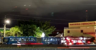 Ônibus azul de linha passando em frente a estabelecimentos comerciais iluminados à noite, sob céu nublado.