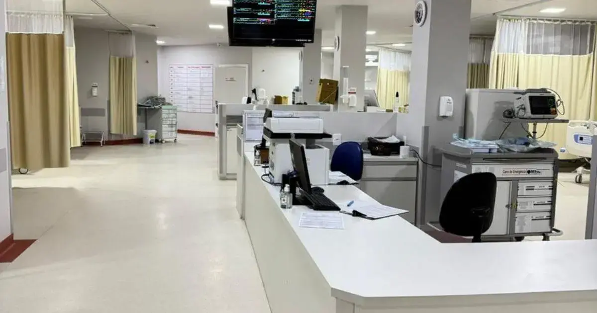 Ambiente interno de um hospital com balcão de atendimento, monitores e cortinas nas áreas de cuidado.