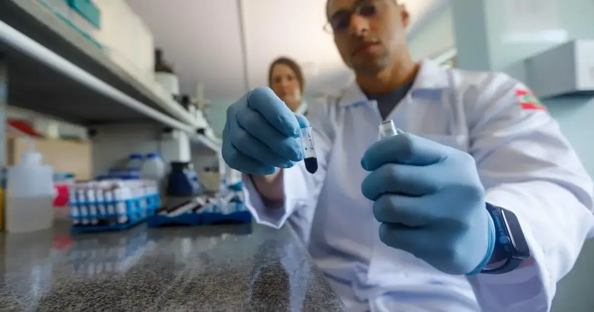 Um profissional de laboratório, usando luvas azuis, segura dois tubos de ensaio enquanto outra pessoa está ao fundo.