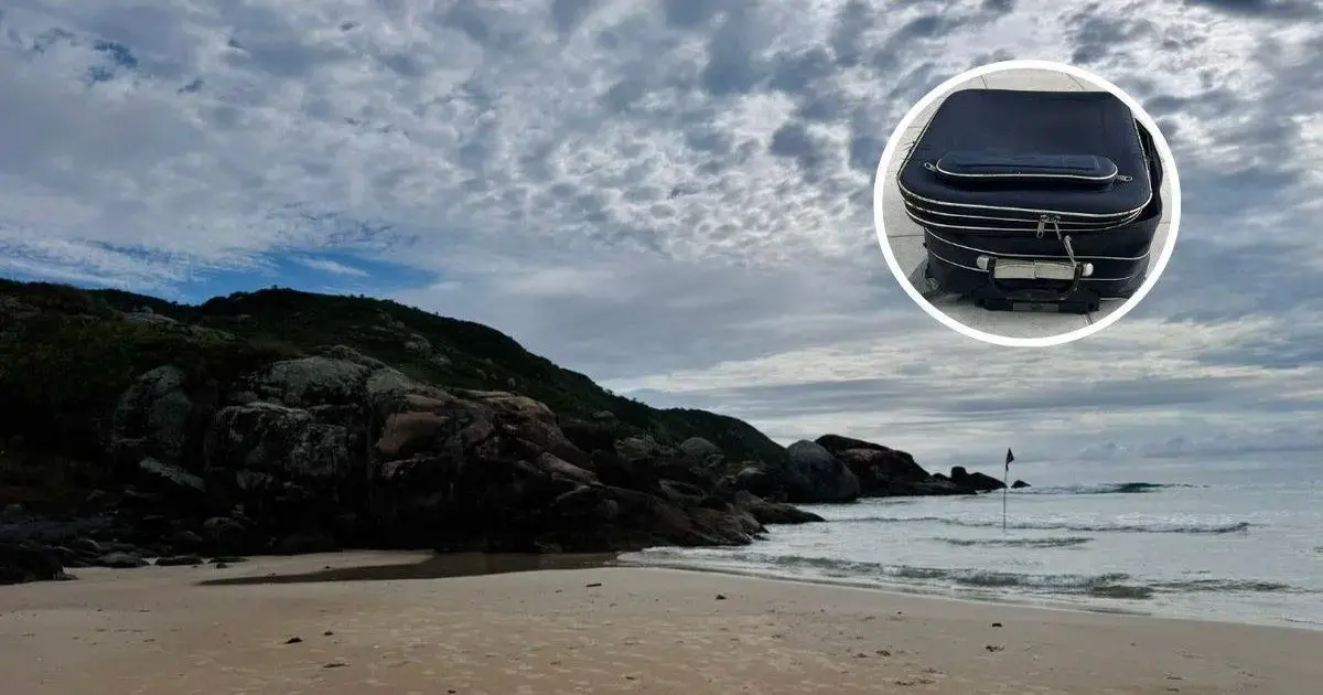 Praia com céu nublado, rochas à beira-mar e uma mala de viagem em destaque.