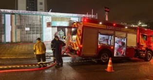 Caminhão dos bombeiros estacionado com funcionários verificando equipamentos em frente a um prédio à noite.