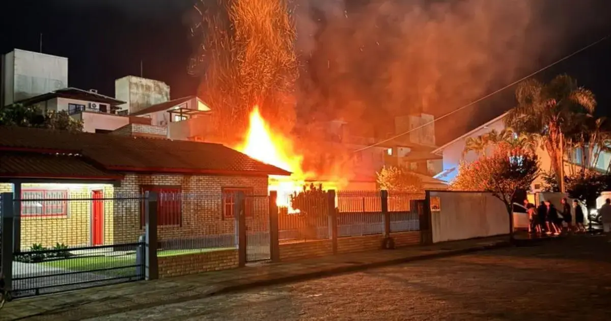 Chamas altas e fumaça em uma casa à noite, com móveis e pessoas visíveis na área externa.