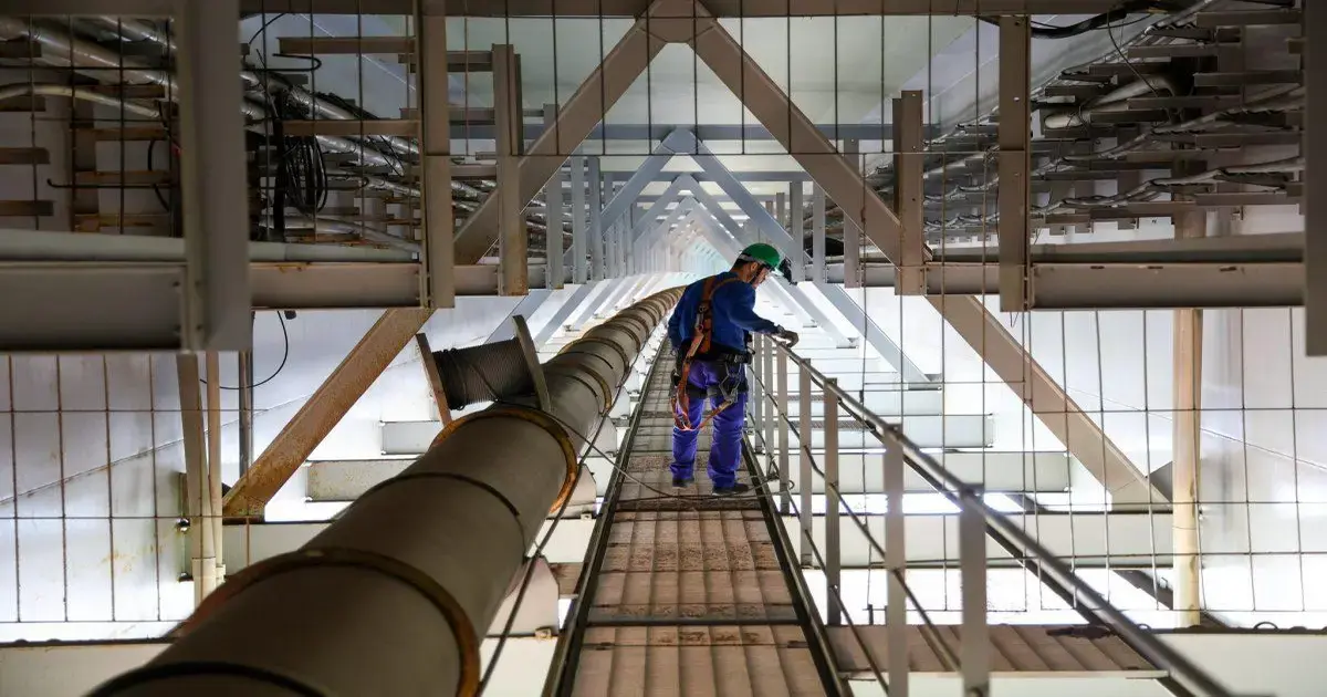 Trabalhador em uma plataforma elevada inspeciona equipamentos sob estruturas metálicas e tubulações expostas.