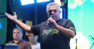 Homem com camiseta preta faz discurso ao microfone, com público ao fundo e iluminação colorida.