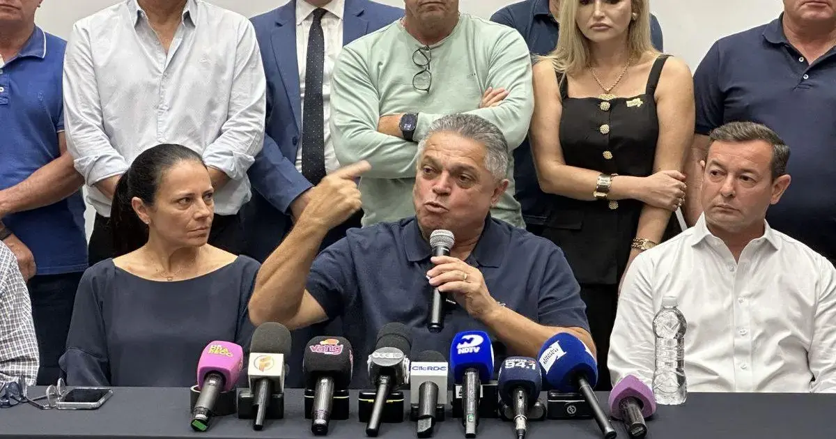 Homem com microfone discute em coletiva, cercado por outras pessoas e microfones em mesa.