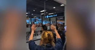 Multidão reunida atrás de uma parede de vidro em um aeroporto, com pessoas acenando e saudações. Placa indicativa "Saída | Exit".