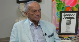 Homem sentado com terno claro, olhando para a frente, com retratos e uma escultura ao fundo. Texto visível: "Senhor Amigo."