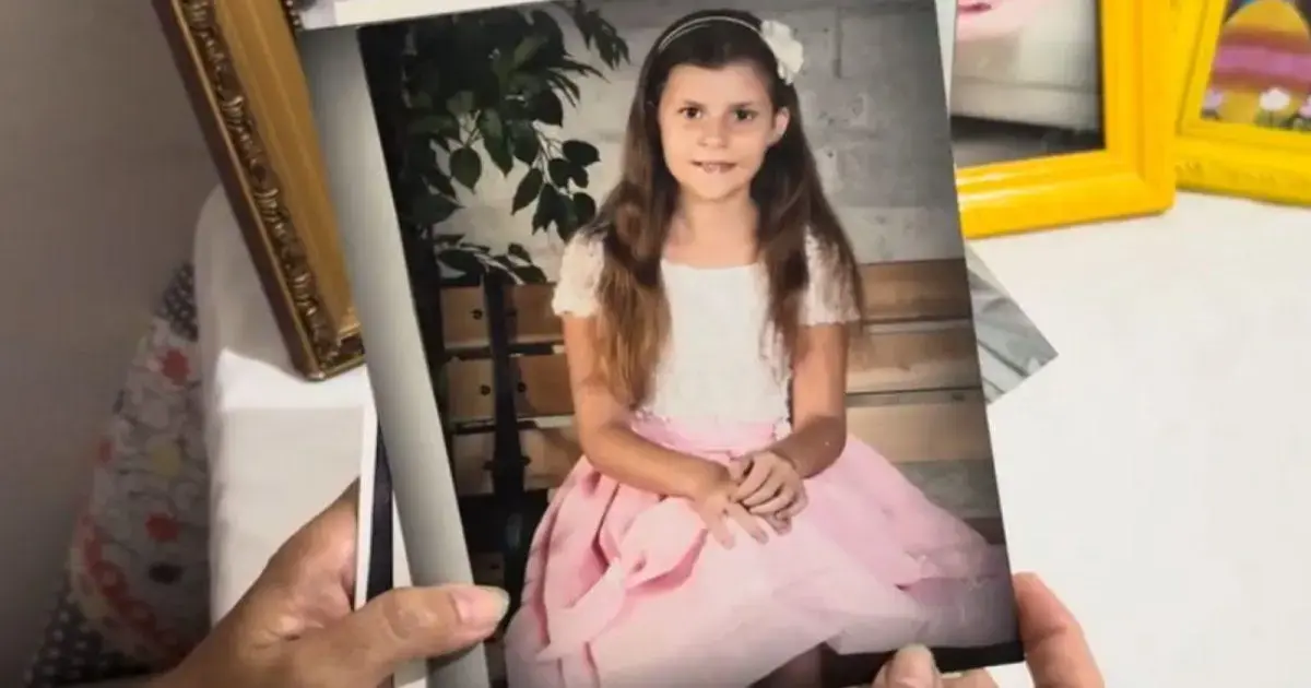 Criança sentada em um banco, usando um vestido rosa, com cabelo solto e um acessório na cabeça.