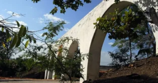 Arcos brancos de um aqueduto, cercados por vegetação, sob um céu azul com algumas nuvens.