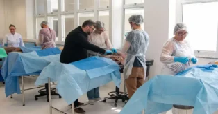 Grupo de pessoas em um laboratório realizando práticas cirúrgicas em corpos simulados cobertos com lençóis.