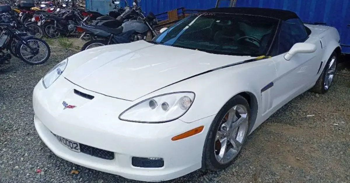 Veículo esportivo branco, modelo conversível, estacionado em área com motos ao fundo.