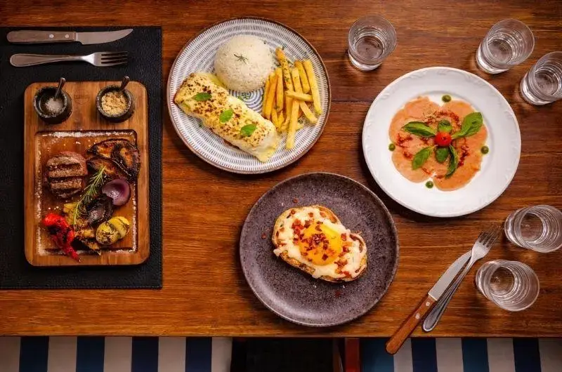 Prato com carnes, legumes grelhados, arroz e fritas, além de pratos adicionais em uma mesa de madeira.