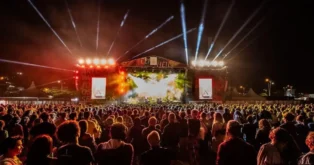 Multidão em um show ao ar livre, com palco iluminado por refletores e fumaça visível.